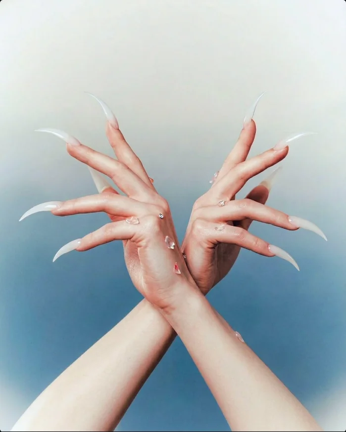 Crossed hands wearing long milky press-on claws with crystal accents against a blue sky
