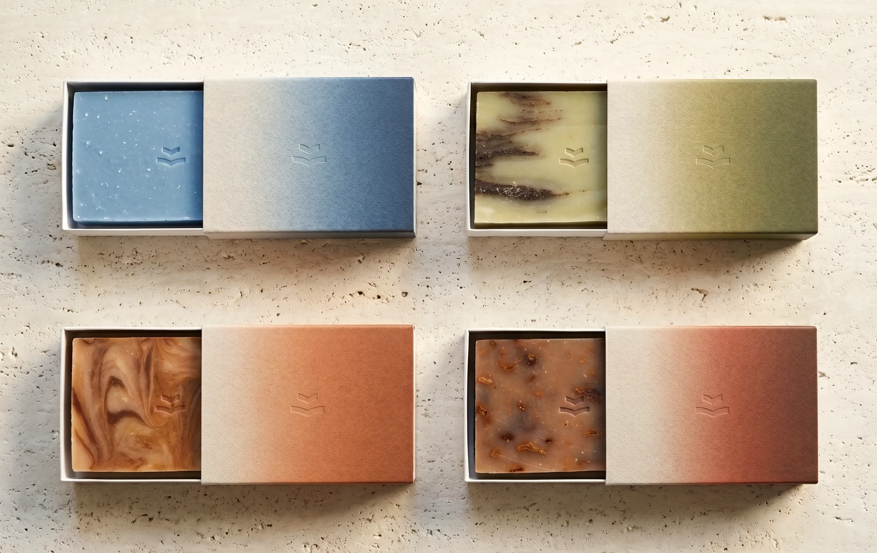 Sage branded soap bars in slide boxes — blue, sage, terracotta, and amber, each embossed with the logomark, on travertine